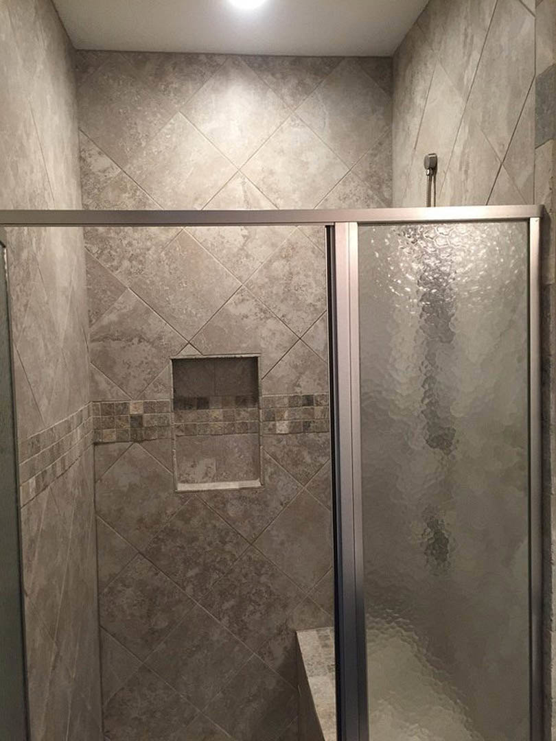 Frameless glass shower door with stone tile walls, white accent wall with sconce, and light gray tile floor
