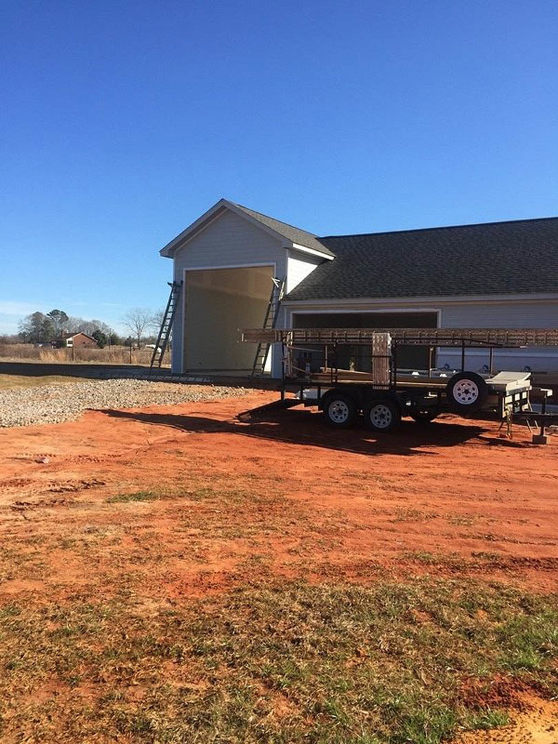 Metal-framed trailer stacked on another trailer parked on dirt in front of a garage, ladders leaning beside garage door, close-up of tire visible, grassy field and sky in