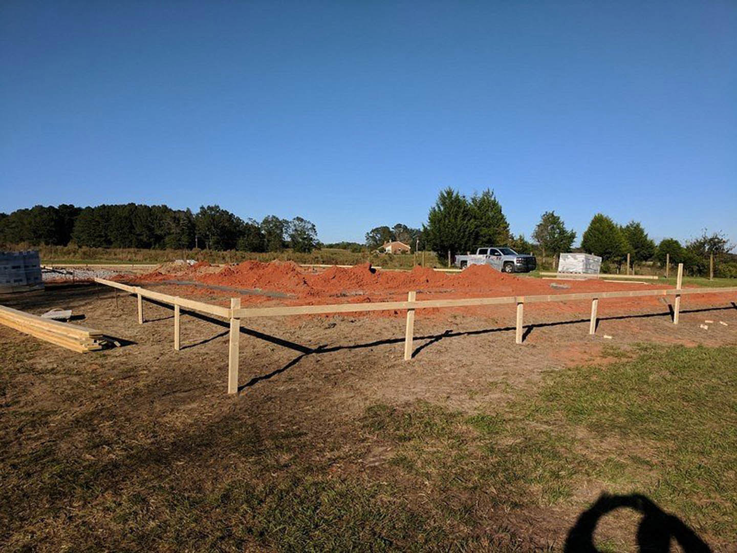 Dirt pile in fenced yard with wooden fence, white truck with black stripes parked nearby, shadow of a hand on ground, blurry building in background, blue sky and trees visible
