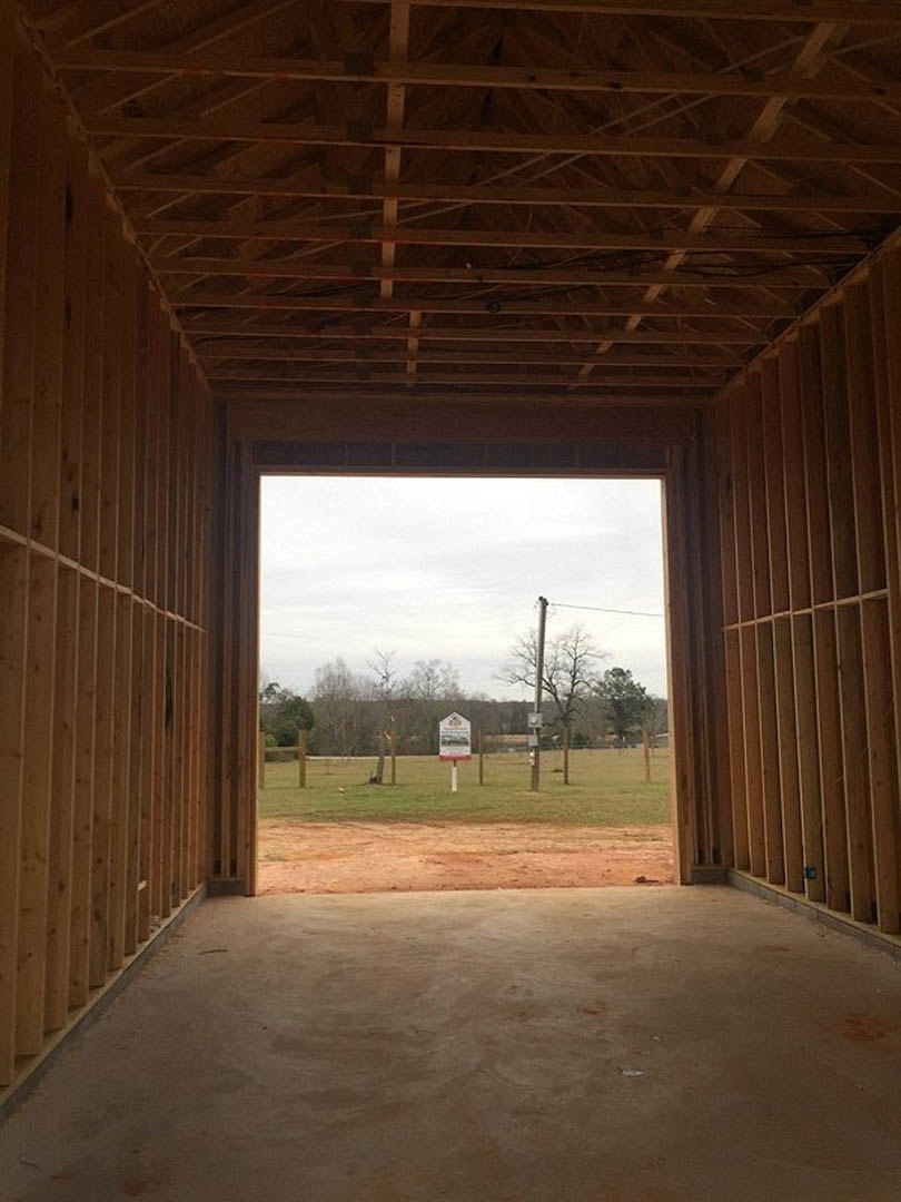Large windows frame a view of grassy field and trees, with wooden beams and shelving visible inside the custom home.