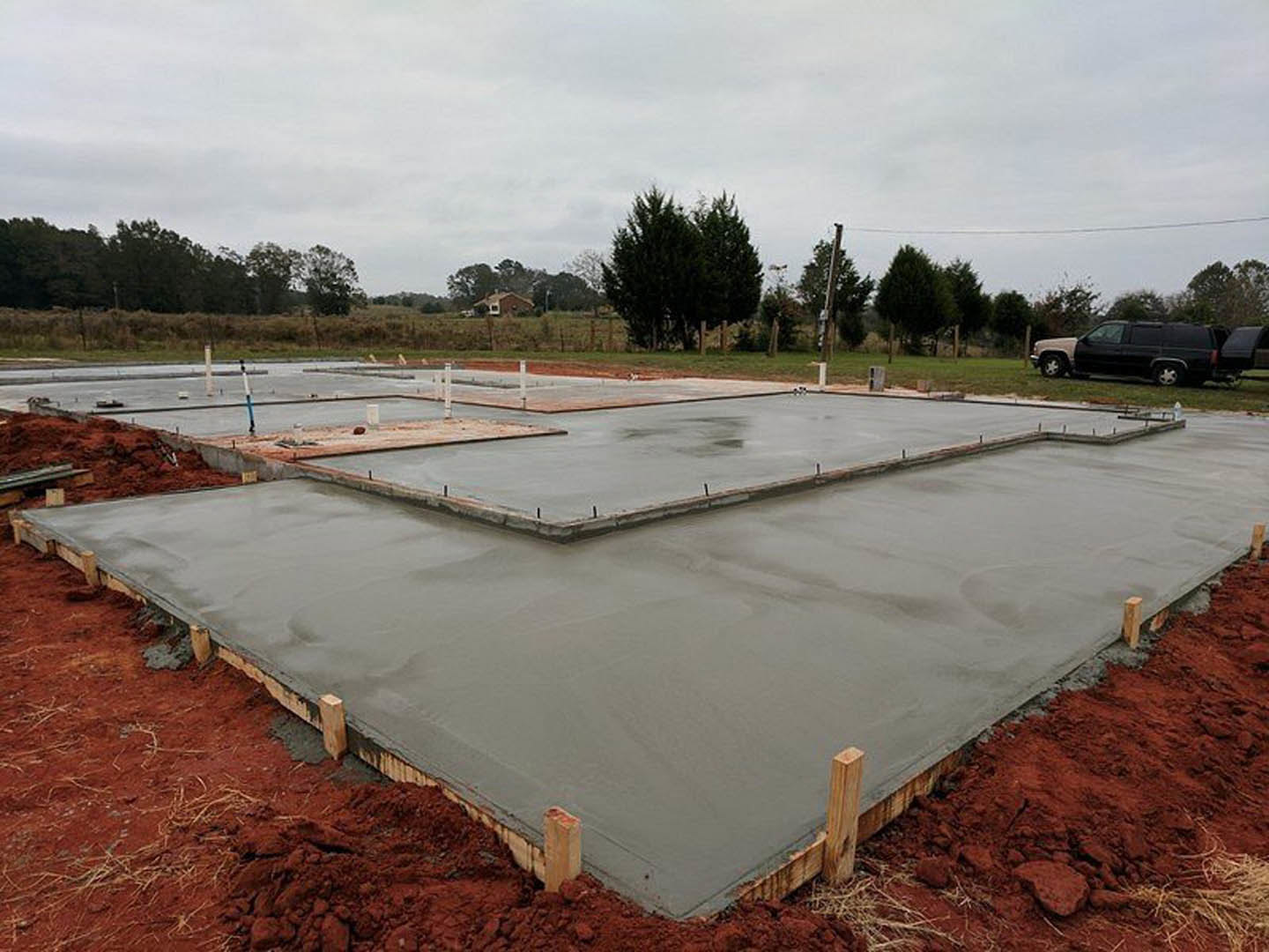 Freshly poured concrete slab surrounded by grassy lawn, black van parked nearby, leafy tree in foreground, distant group of trees, cloudy sky overhead, partial view of a person and