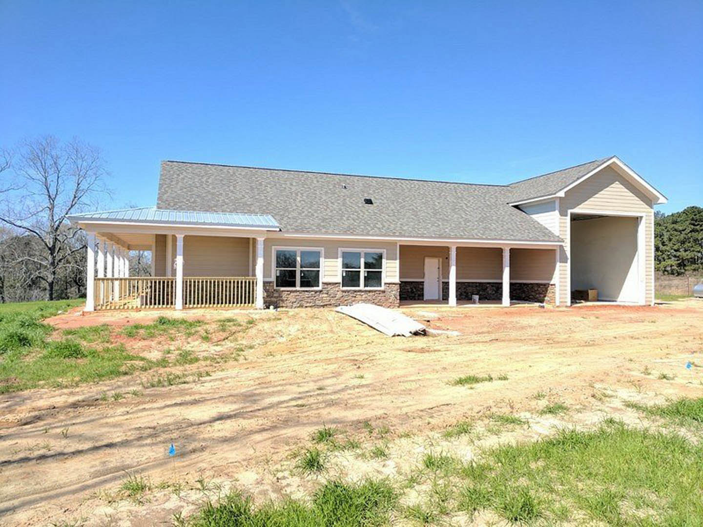 Stone and brick house under construction with exposed wood planks, metal railing, leafless tree, dirt road, and white object on ground