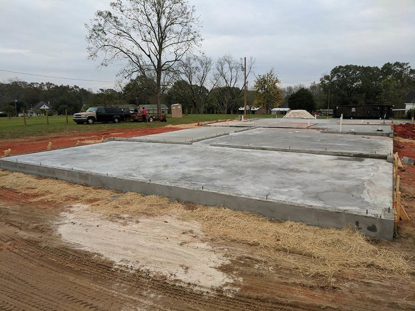 Concrete foundation slab with exposed metal rods, black pickup truck parked nearby, dirt road with tire tracks, leafless trees and grassy land under cloudy sky