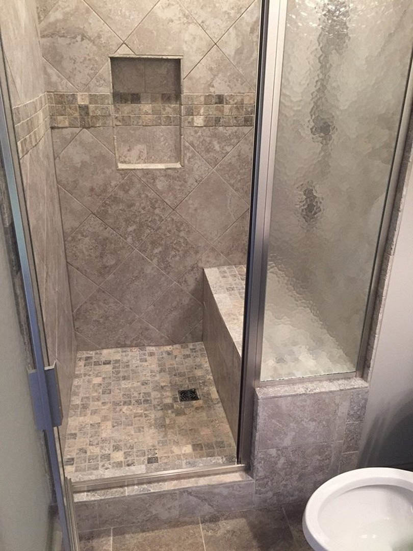 Frameless glass shower door with stone tile walls and floor, adjacent white toilet, bathroom mirror partially visible