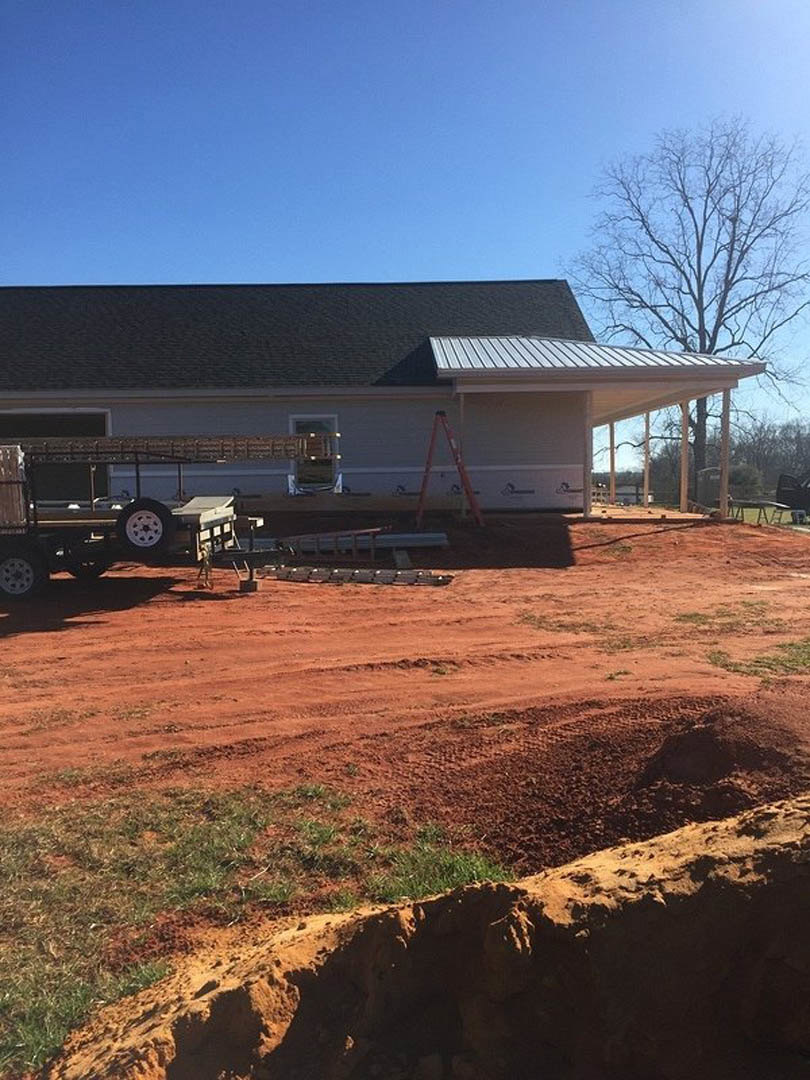 Partially built home with exposed framing, red ladder leaning against exterior, leafless tree nearby, construction trailer parked on dirt ground