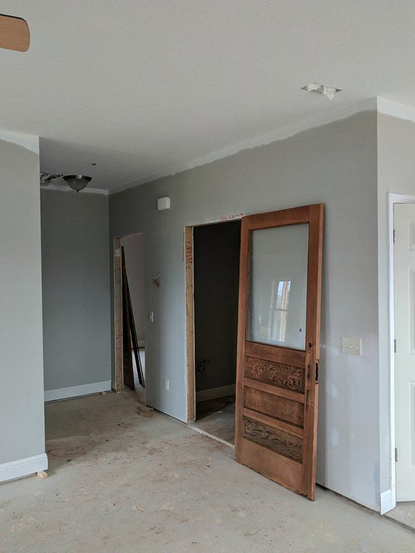 Wooden door with glass window partially open, white wall with three-knob switch, black accent wall with wooden frame, white door hinge visible, light plaster walls and flooring.