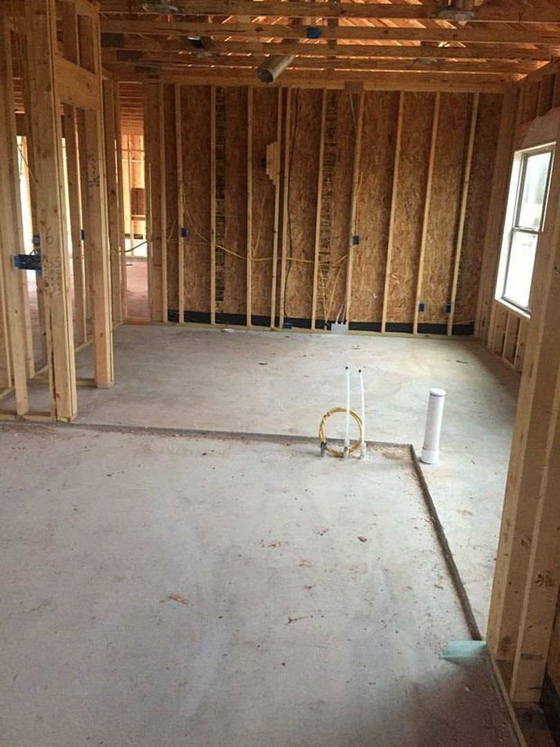 Framed wooden wall with exposed beams, large window letting in natural light, unfinished concrete floor with yellow tubing, blue construction material on wood structure