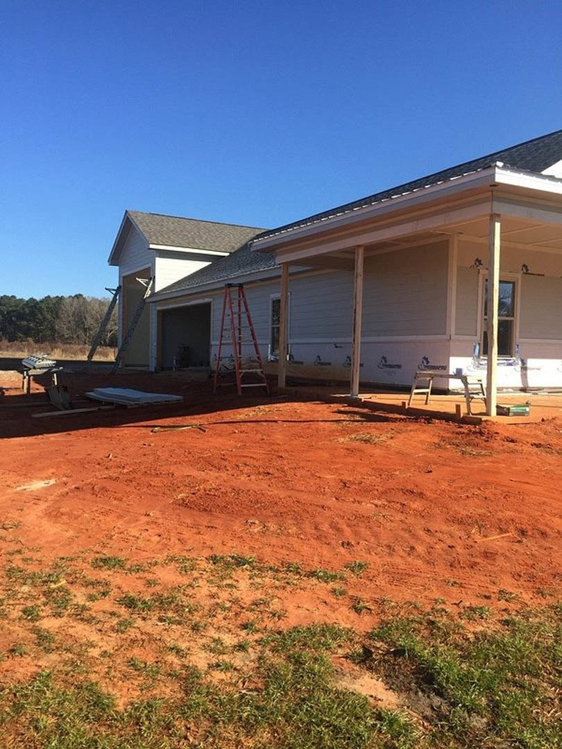 Partially built house with exposed framing, red dirt yard, white exterior walls, black door with white trim, porch area, and ladder leaning against garage.