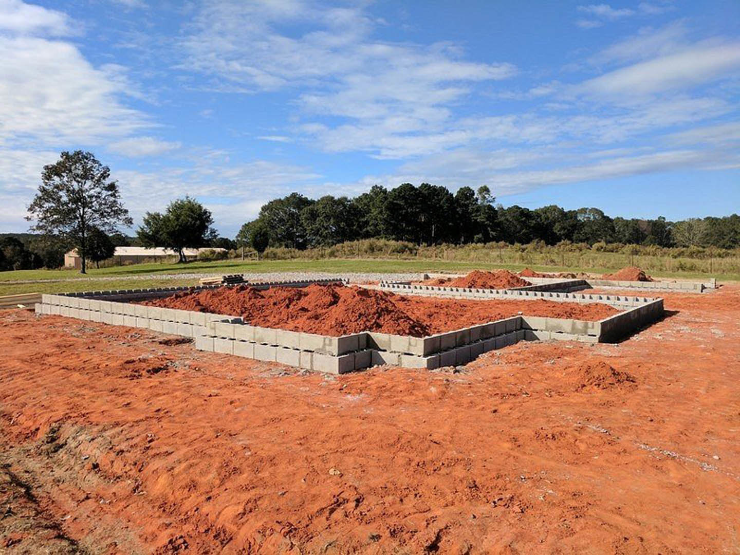 Red dirt construction site with scattered bricks, open field, mature tree, and blue sky with clouds in the background