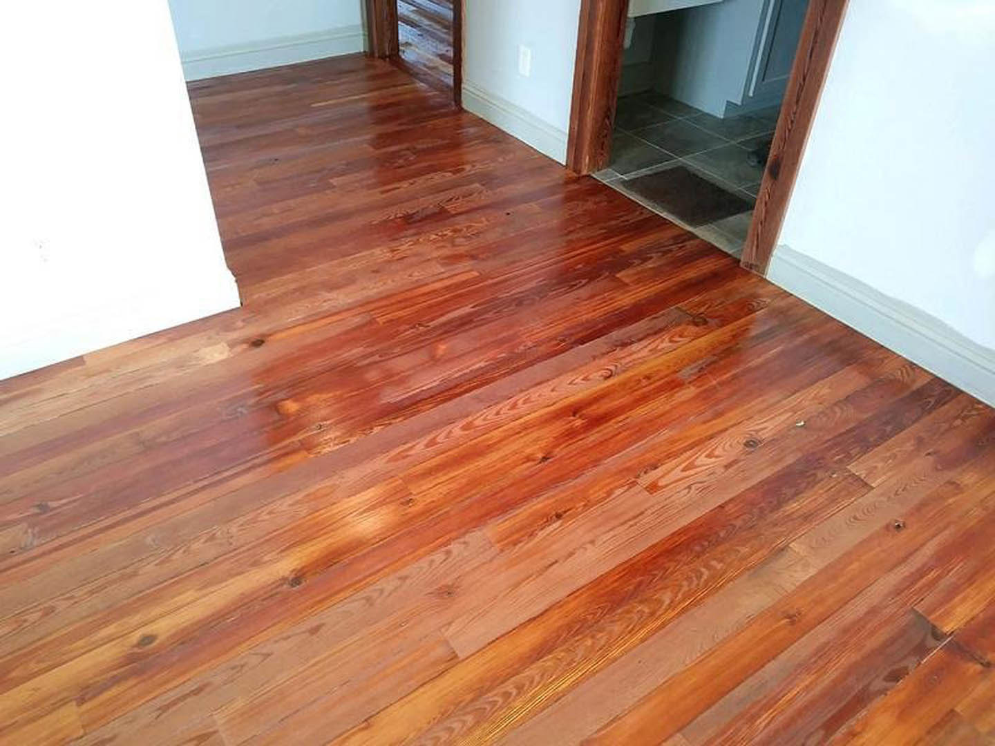 Hardwood plank flooring in a bright room with white walls, open doorway leading to kitchen, large window, and wooden trim