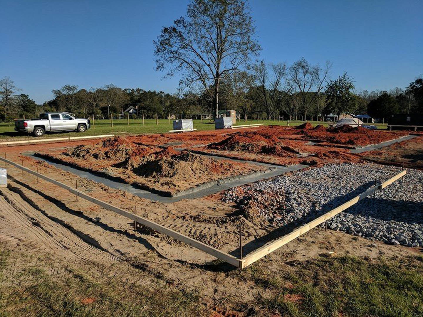 Dirt and gravel construction site with a white truck parked on grass, leafless tree, wooden beam on rocky ground, and blurred figure in foreground