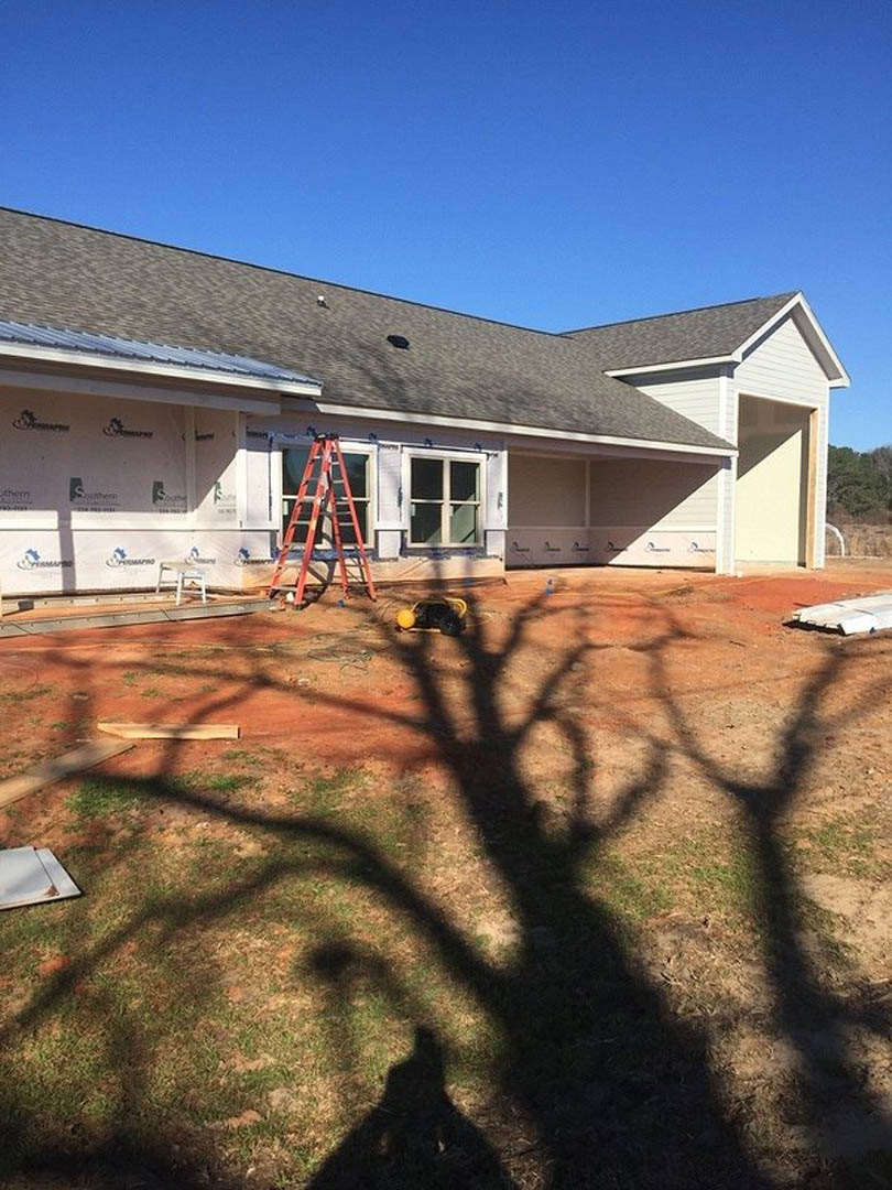 Partially built house with exposed framing, ladder leaning against large window opening, tree shadows cast on ground, construction materials scattered nearby