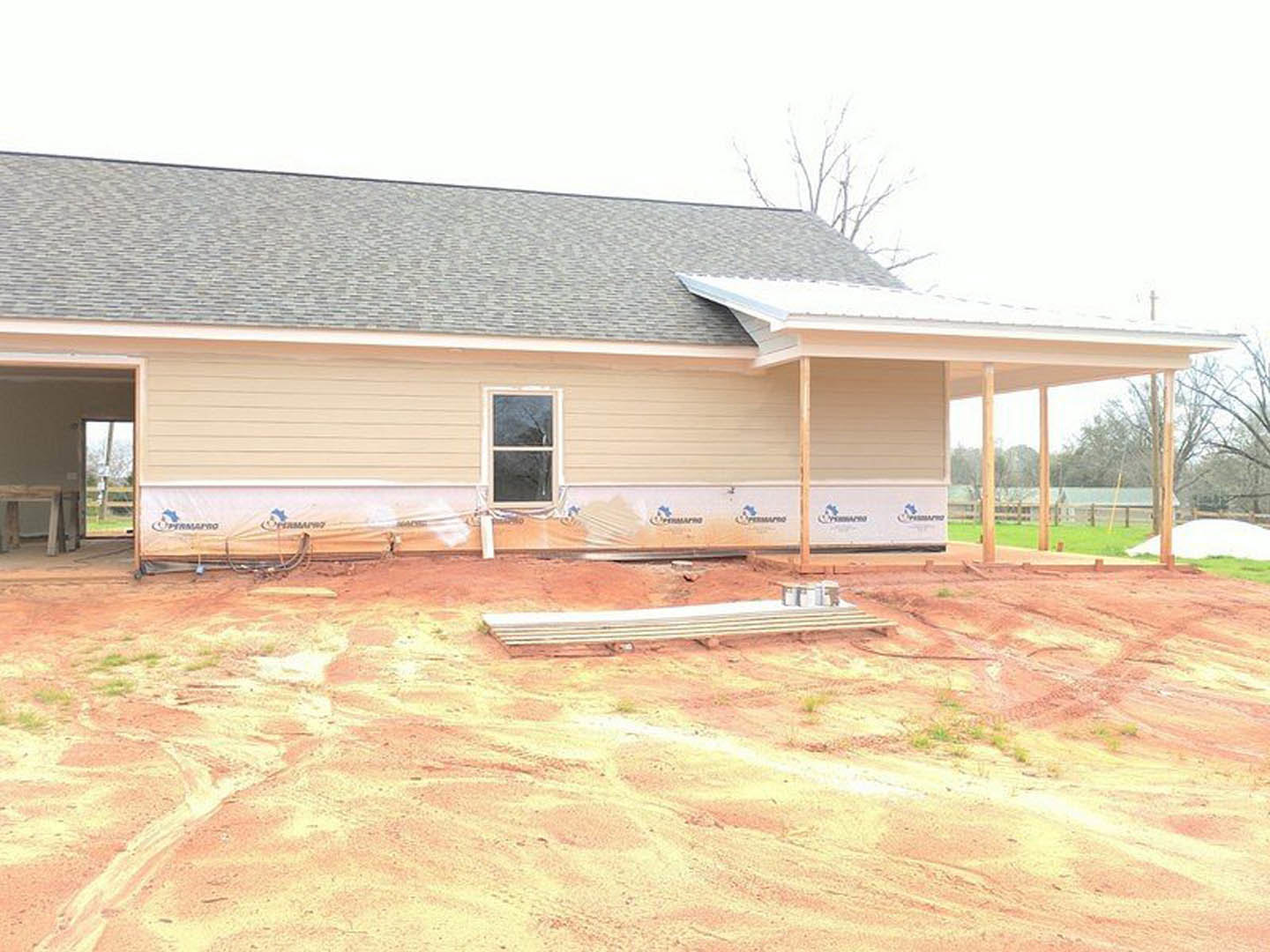 Partially built house with exposed framing and roof, white-framed window installed, metal planks and construction materials scattered on dirt ground, cloudy sky overhead