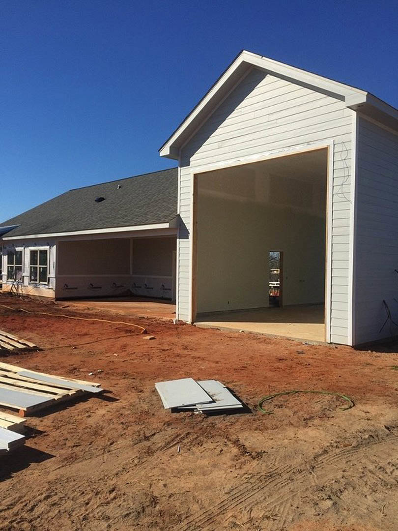 White siding house under construction with open garage door, exposed framing, dirt yard, and stacked wooden pallets in foreground