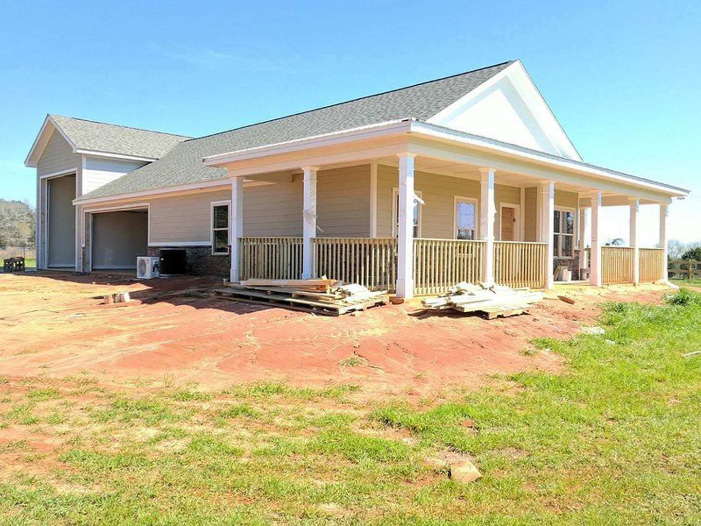 Two-story house under construction with white siding, covered porch supported by white pillars, round attic vent, pile of lumber near wooden fence, grassy yard, and multiple