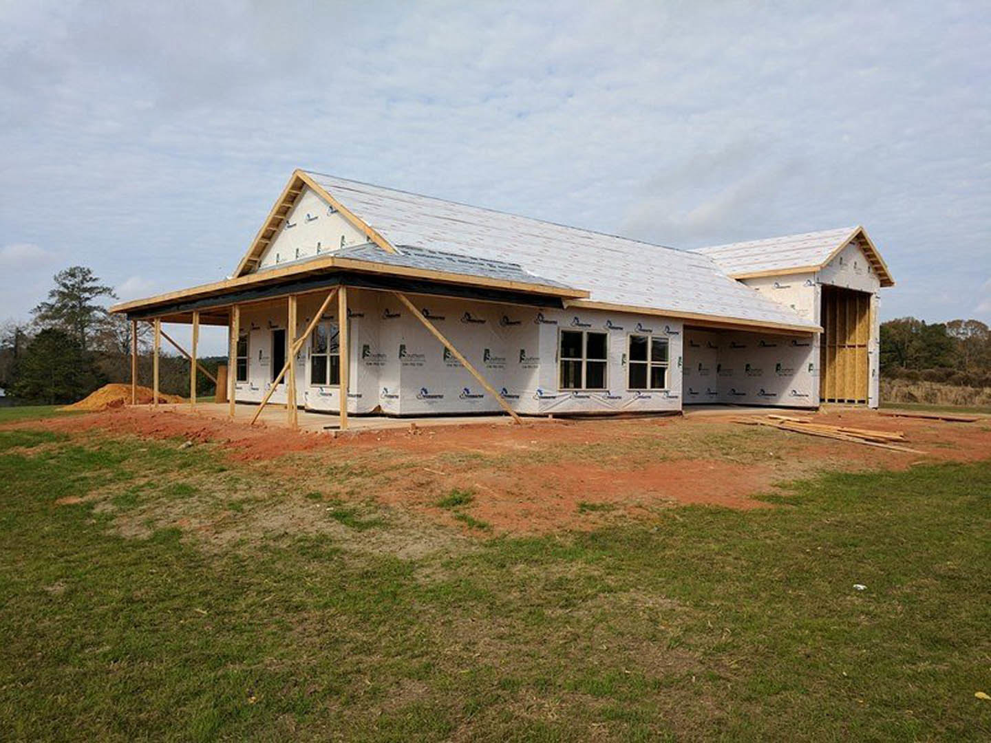 Partially built house with exposed wooden framing, unfinished roof, grassy yard, scattered construction materials, leafy tree, and clear blue sky