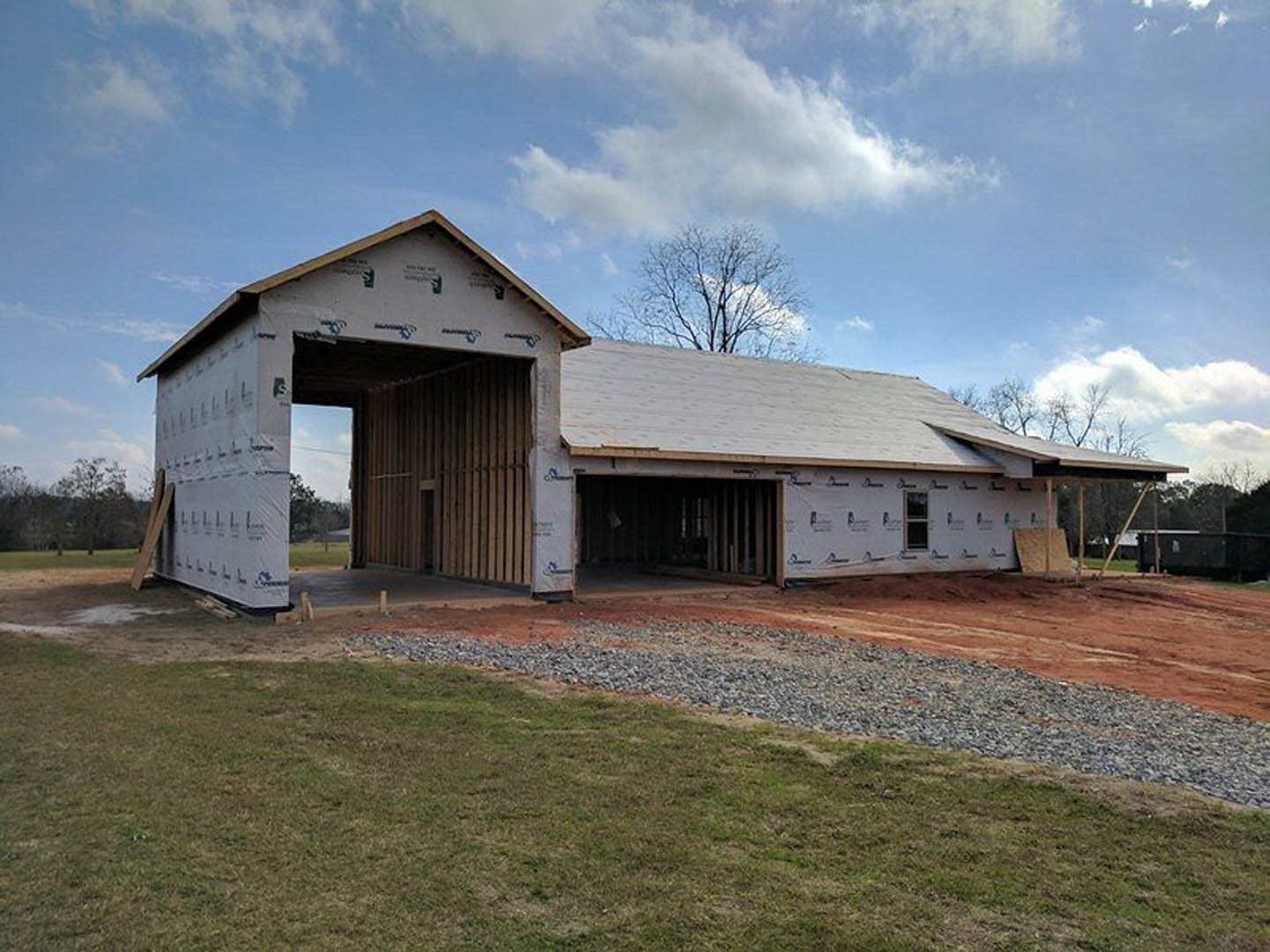 Partially built house with exposed wooden framing, large open doorway, gravel and grass foreground, tree and cloudy sky in background