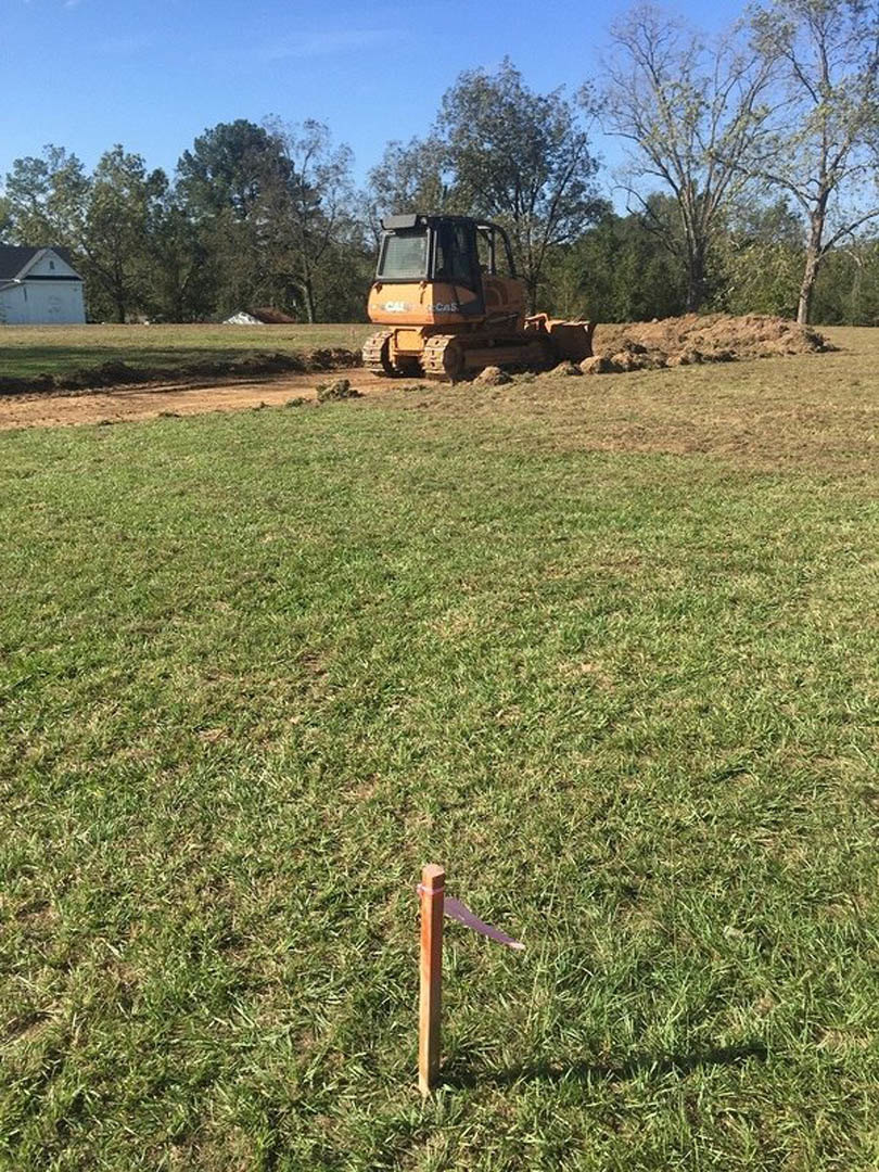 Yellow bulldozer parked on dirt lot surrounded by grassy field, leafless trees in background under blue sky