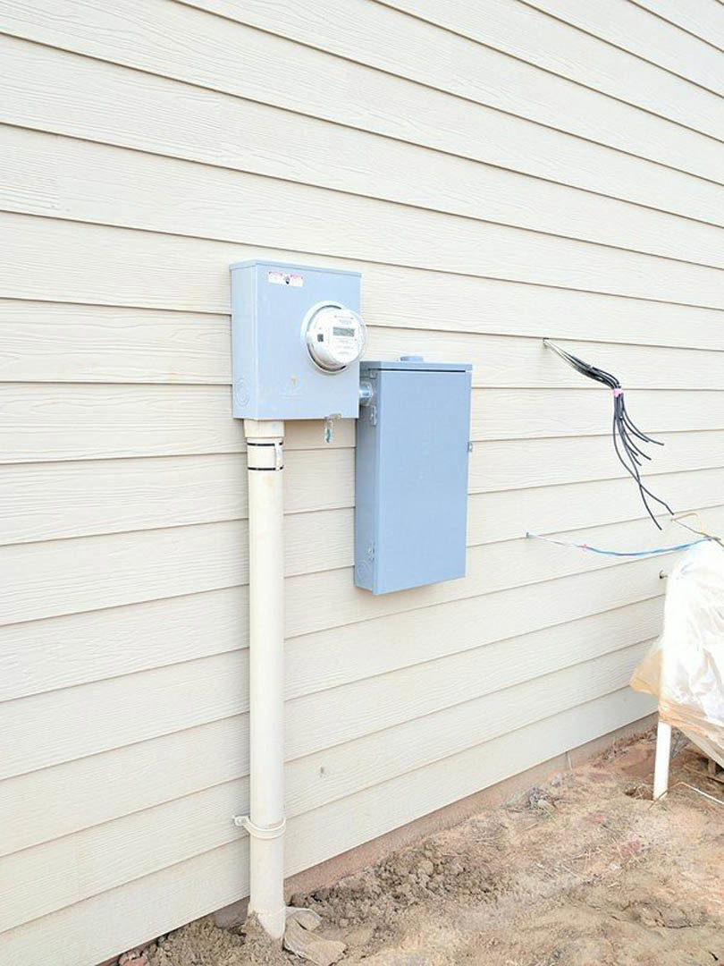 White electrical meter box mounted on exterior wall with visible conduit pipe and wiring, surrounded by white siding.
