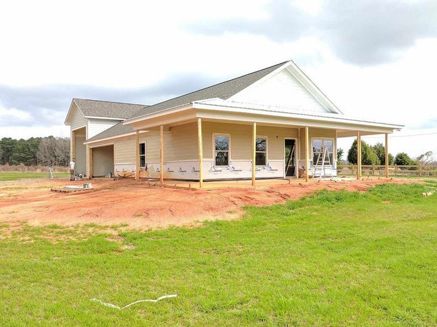Partially built house with exposed wooden framing, covered porch, yellow-trimmed windows, green lawn with a white marking, and trees visible through windows under a cloudy sky.