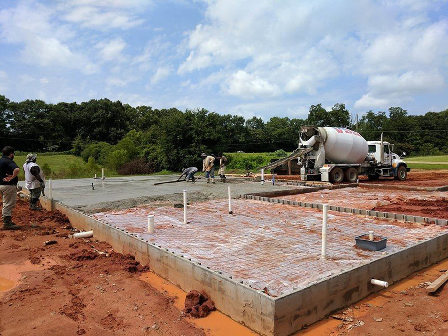 Concrete foundation under construction with workers, cement mixer truck, and scattered building materials; blue sky with clouds overhead.
