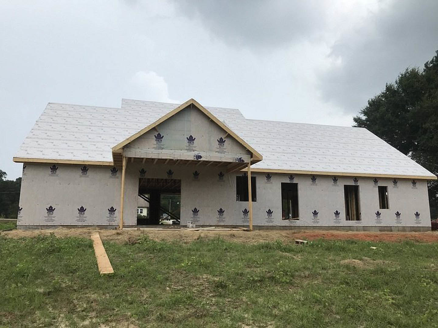 Partially built house with exposed framing and roof, surrounded by grass and mature trees under a cloudy sky