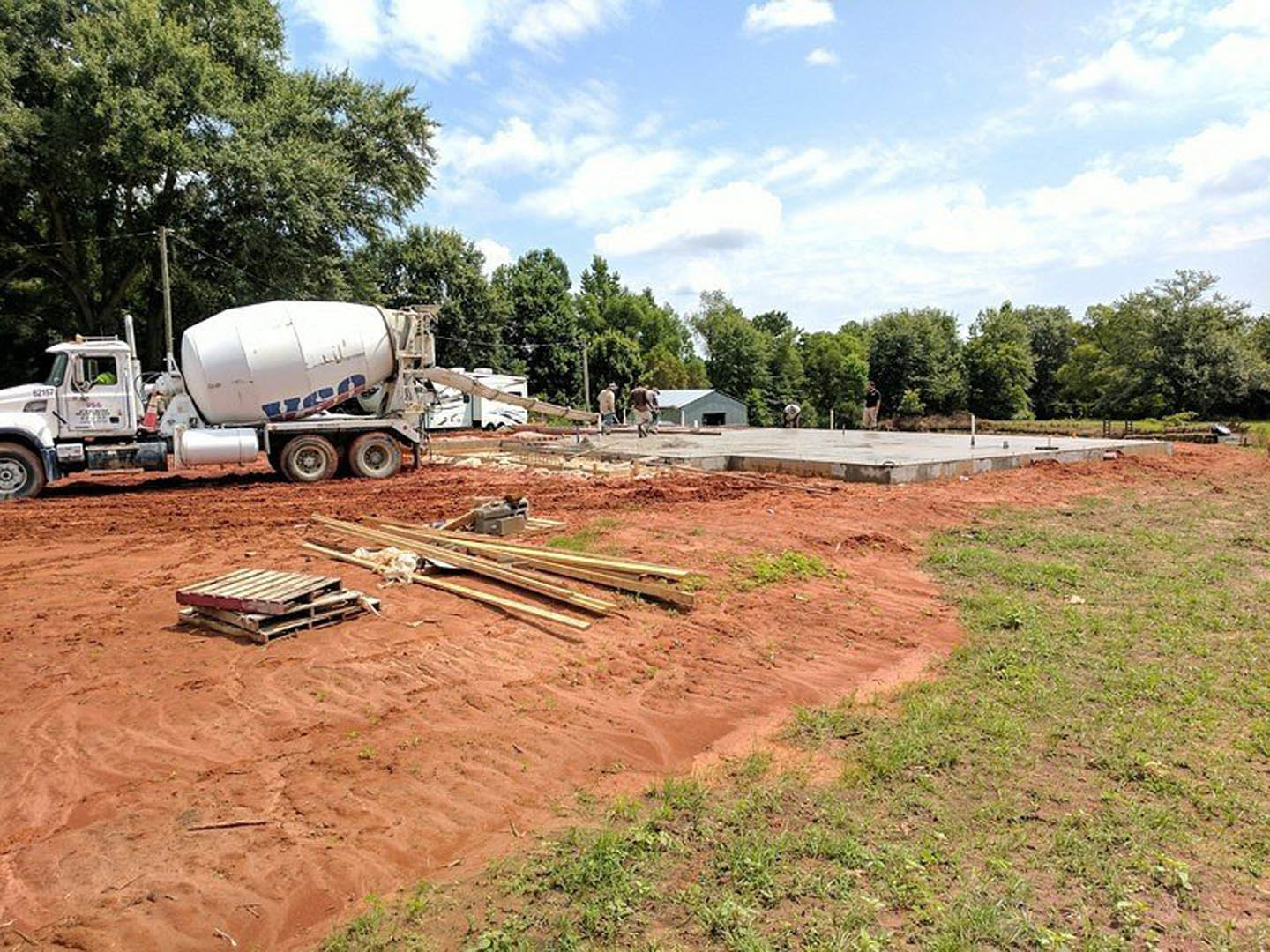 White concrete mixer truck parked on red dirt road, stack of wooden pallets nearby, blue sky with scattered clouds and trees in background.