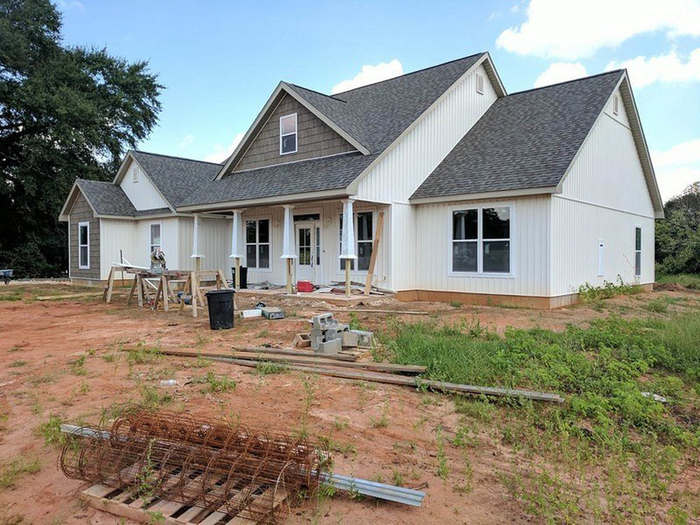 Partially built house framed with wood, surrounded by a wire fence, black trash can near construction area, tree and Robert Frost Farm visible in the background under a blue sky