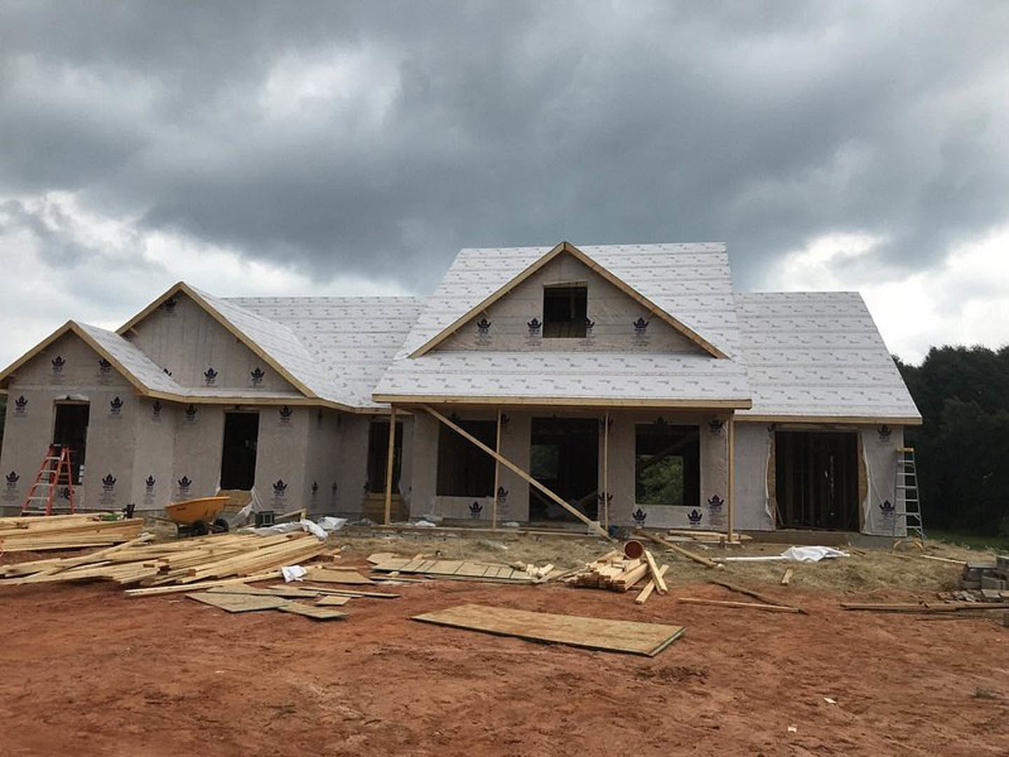 Framed wooden structure of a house under construction, exposed beams and plywood, dirt ground, cloudy sky overhead
