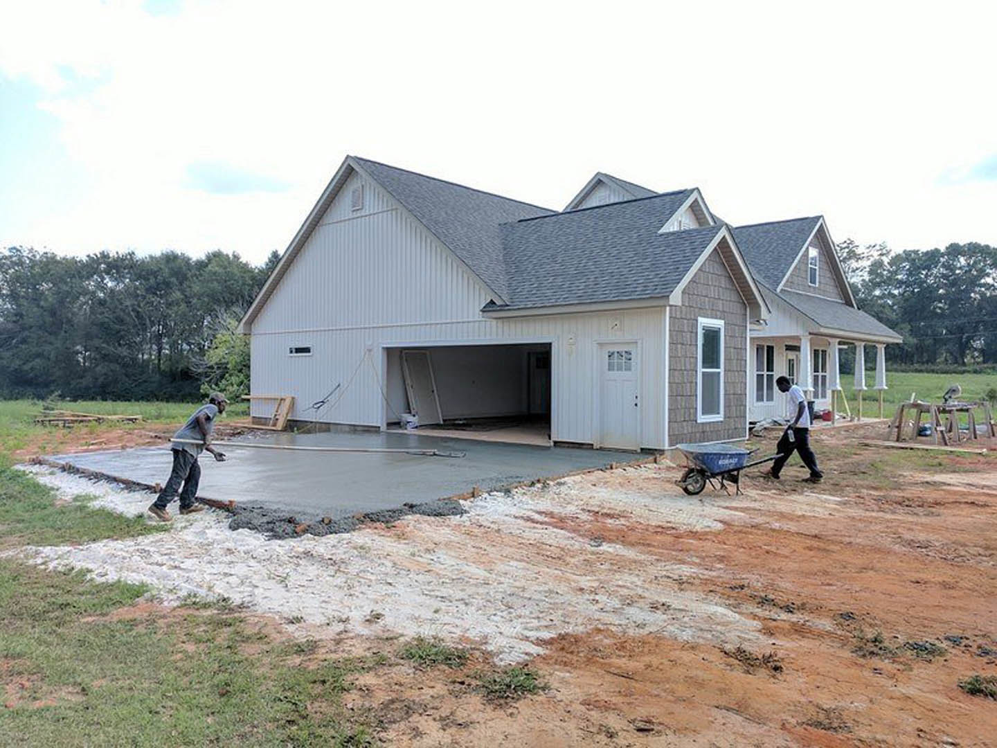 Two men working on a freshly poured concrete slab in front of a house with attached garage, surrounded by trees and cloudy sky
