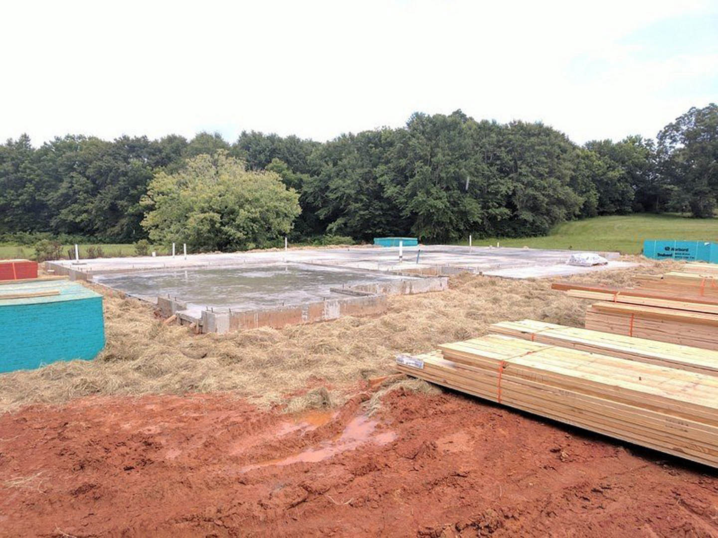 Stack of lumber on dirt construction site, blue tarp covering materials, leafy trees in background, unfinished ground.