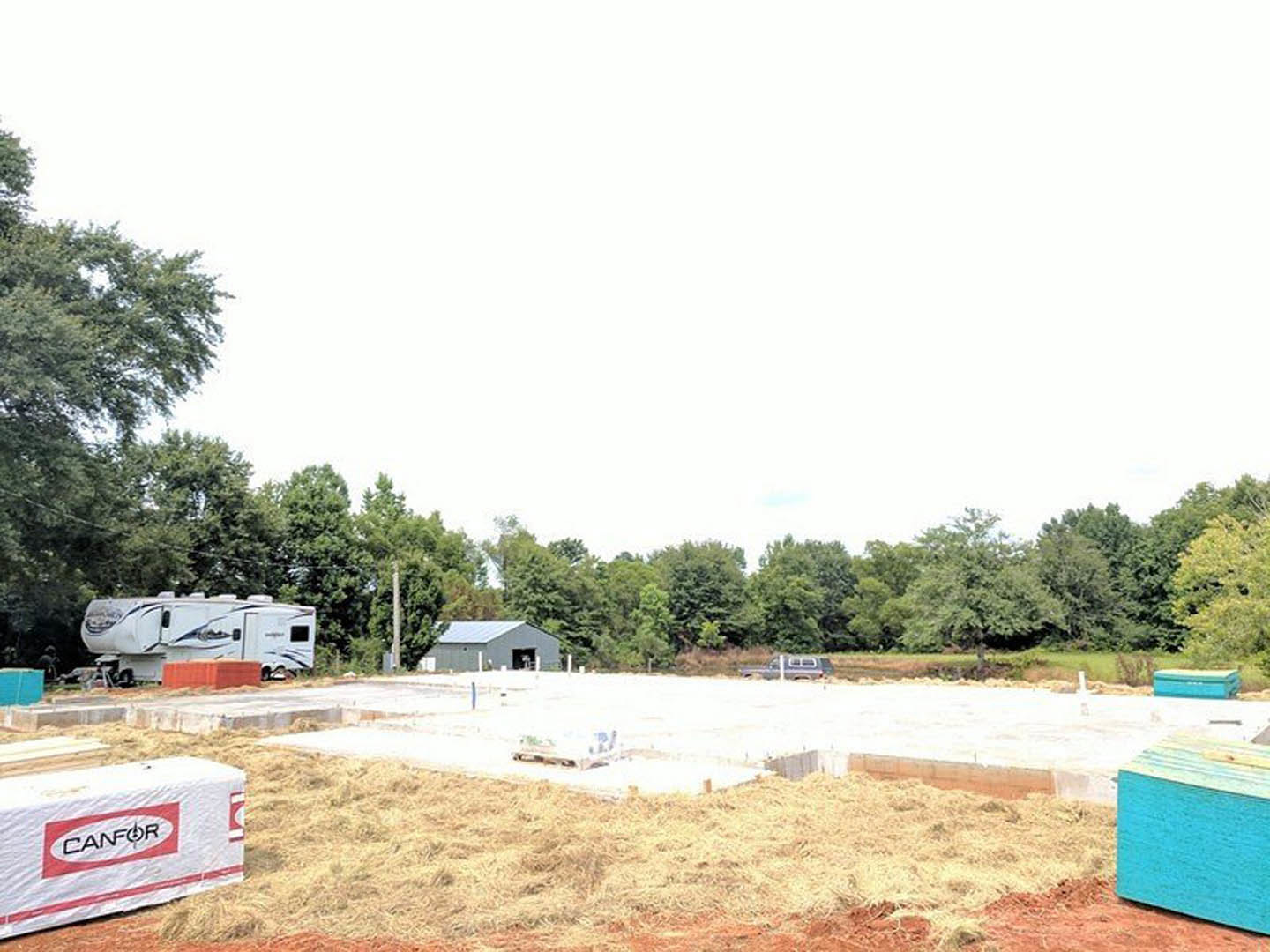 Construction site with white and red trailers, open building door, blue construction materials, waste container, and surrounding trees under a clear sky