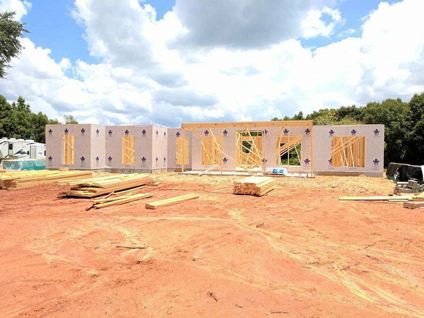 Wood-framed house under construction with exposed beams, piles of lumber on dirt ground, cloudy sky overhead
