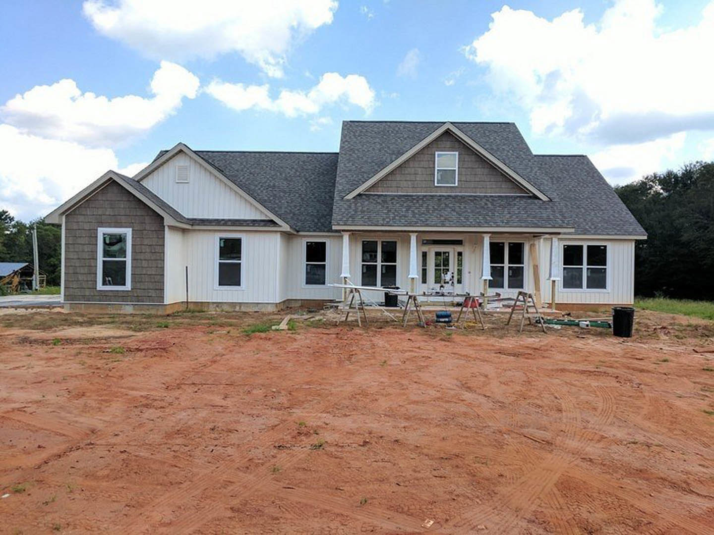 Brick house under construction with white roof, white-framed window, dirt road in foreground, ladder leaning against exterior, black trash can on wooden surface, scattered