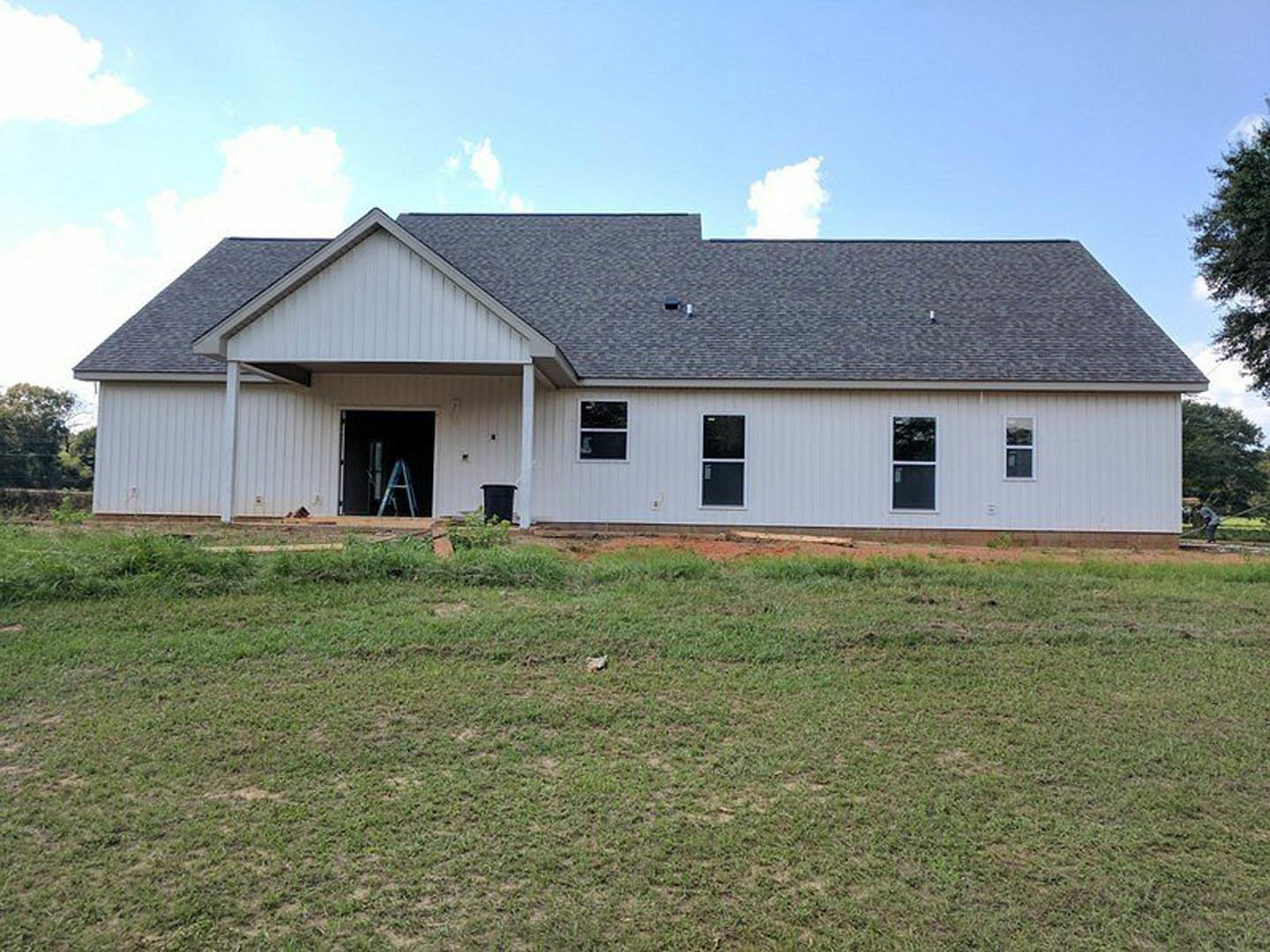 Two-story farmhouse under construction with white-framed windows, covered porch, brick chimney, and ladder in doorway, surrounded by green grass under cloudy sky