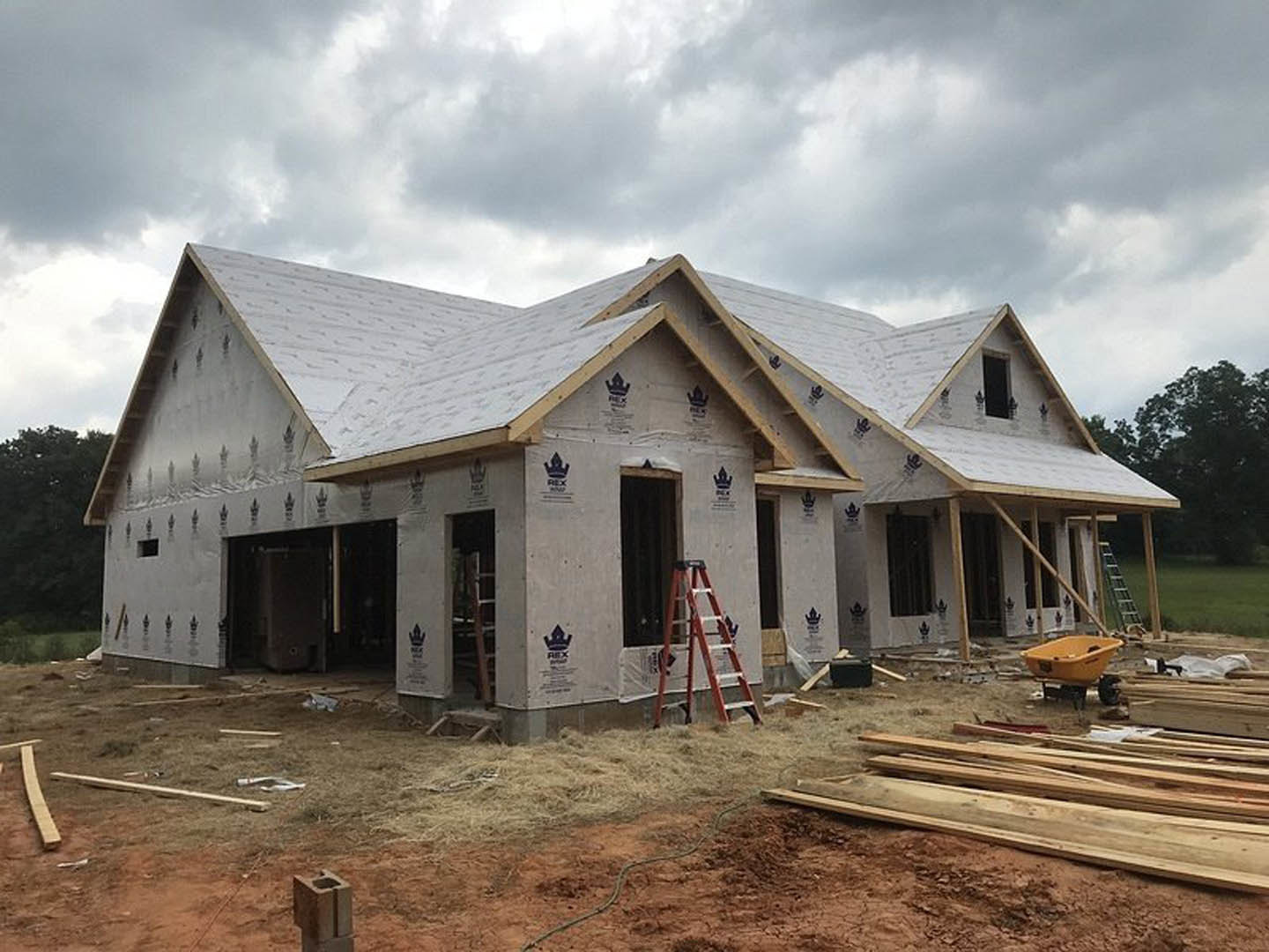 Partially built house with exposed framing, black door with red handle, ladder leaning against exterior wall, pile of lumber on ground, yellow plastic tub on cart, cloudy sky