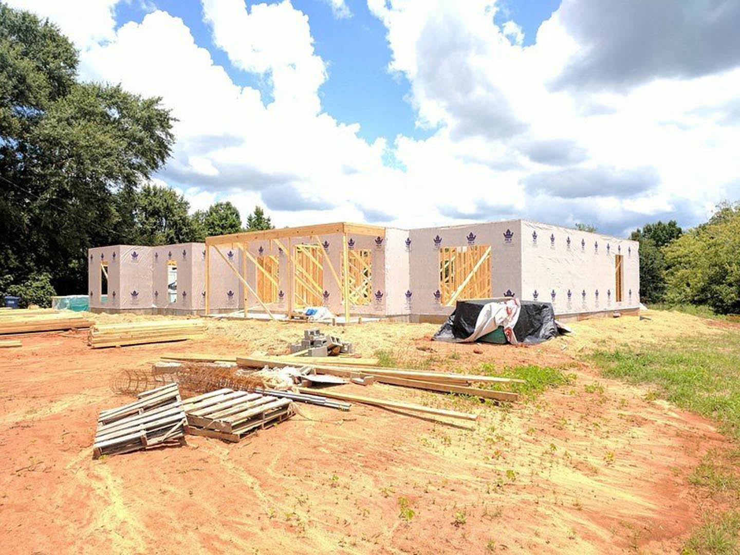 Wood-framed house under construction with exposed beams, scattered wood planks, metal bars, and a person lying on a green container; blue sky with clouds and grassy lot in