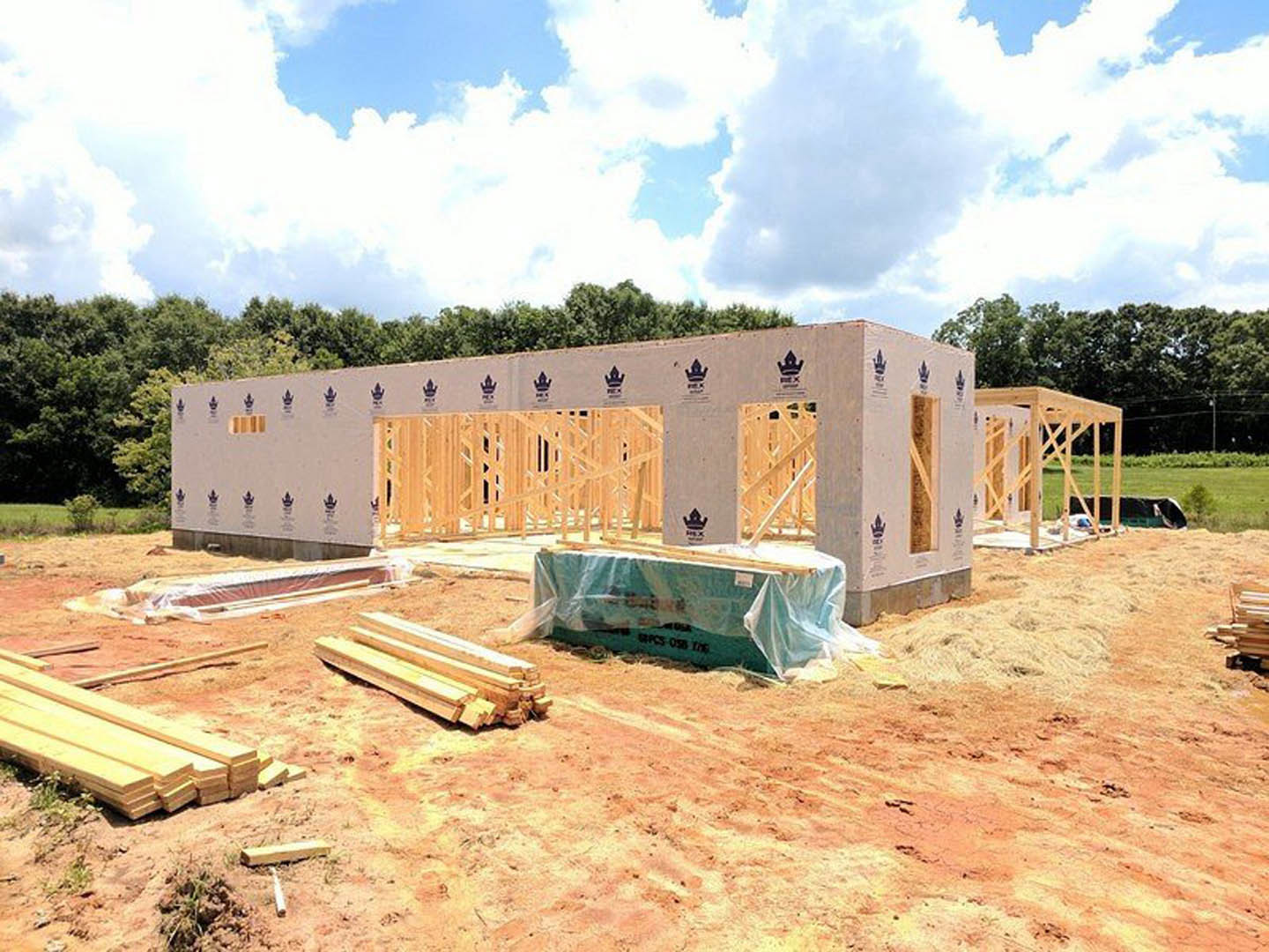 Wood-framed house under construction with exposed beams, blue tarp covering part of the structure, piles of lumber and plastic-wrapped materials on the ground, surrounded by trees