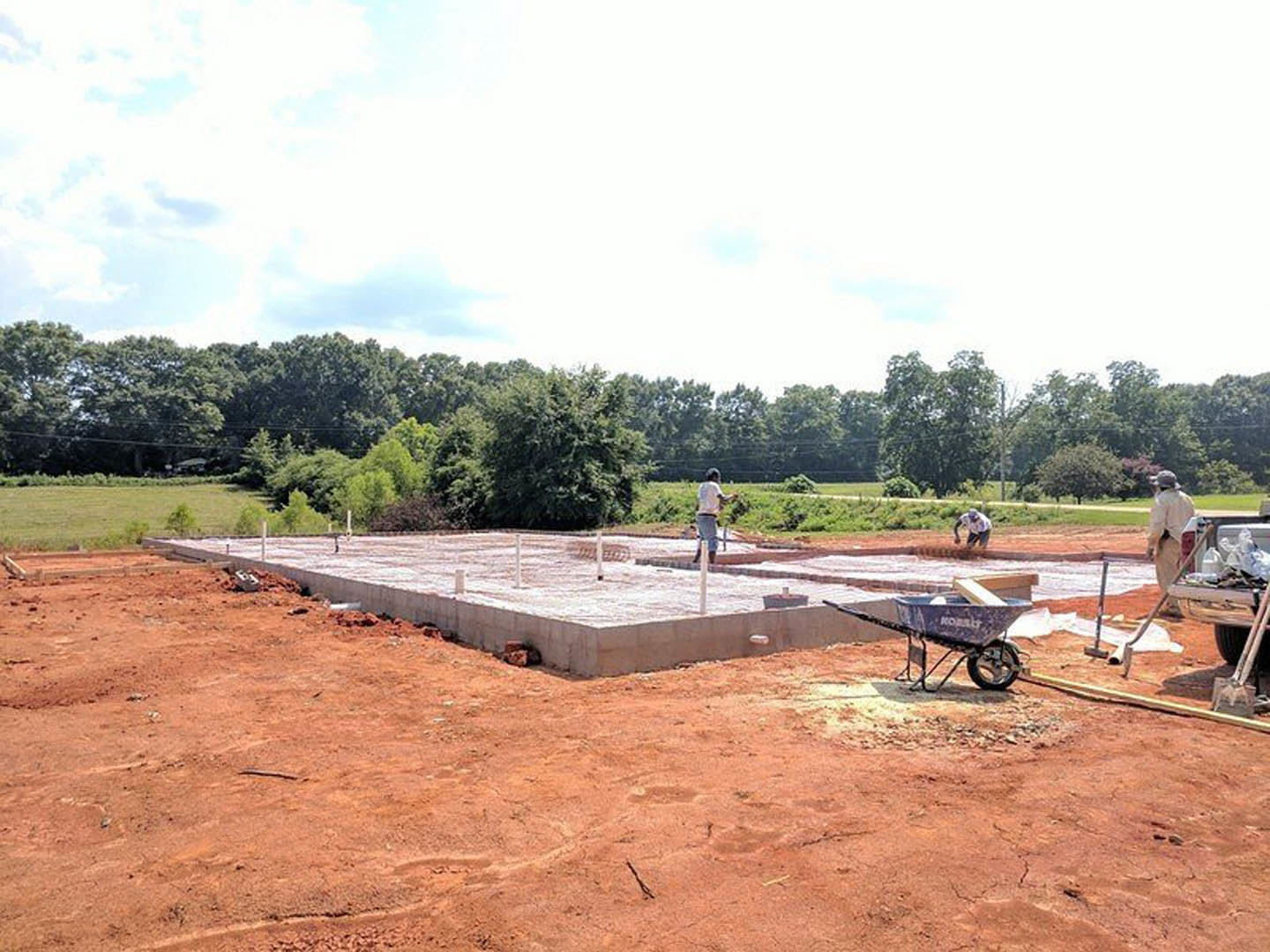Workers pouring concrete foundation on construction site, surrounded by soil, grass, and scattered tools; wheelbarrow with blue wheel in foreground, trees and cloudy sky in