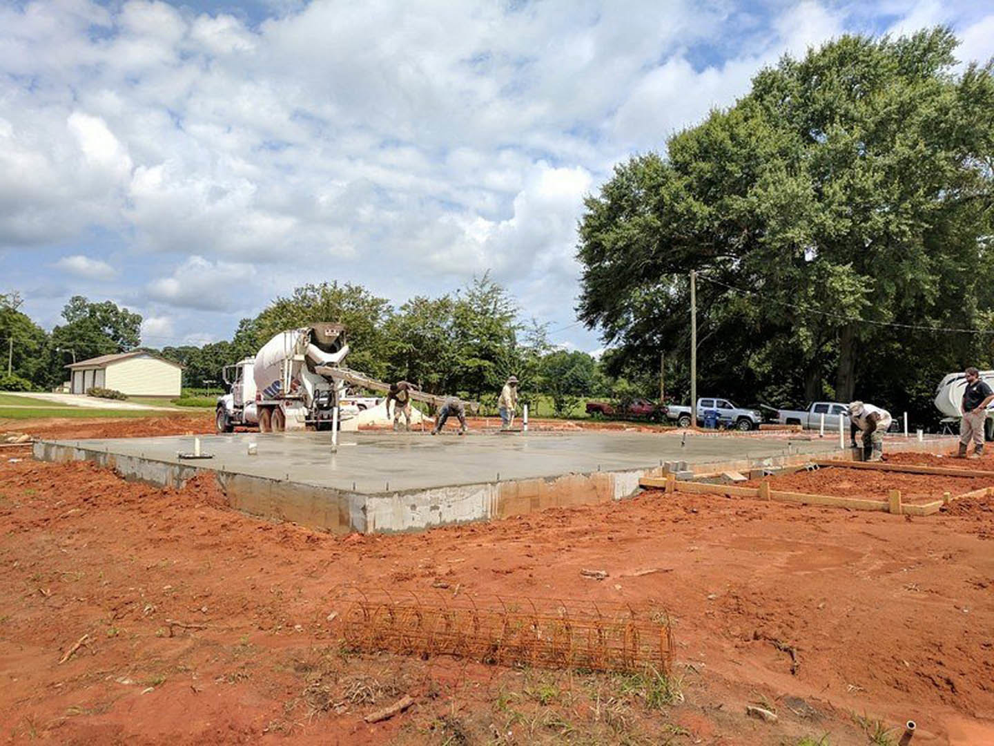 Concrete foundation poured at residential construction site, concrete truck parked nearby, workers in black shirts standing on dirt ground, white house with brown roof in