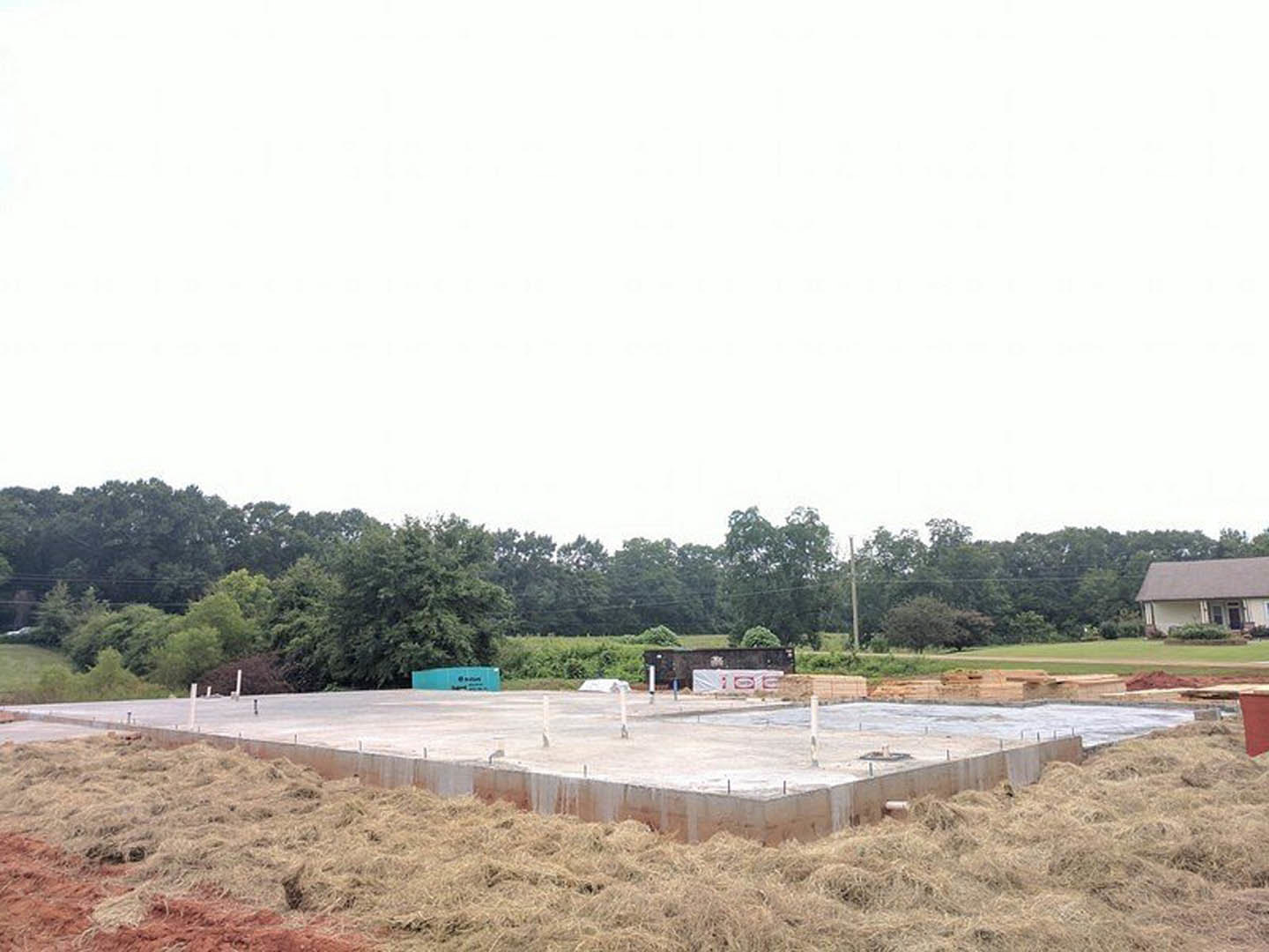Concrete foundation surrounded by grassy field, scattered trees in background, open doorway visible on unfinished structure, blue sky overhead