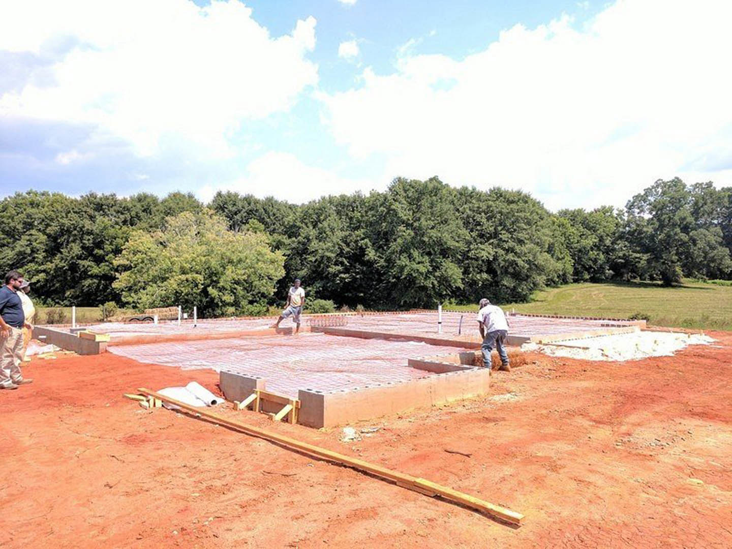 Concrete foundation under construction with workers, exposed soil, scattered building materials, and surrounding trees beneath a blue sky with clouds