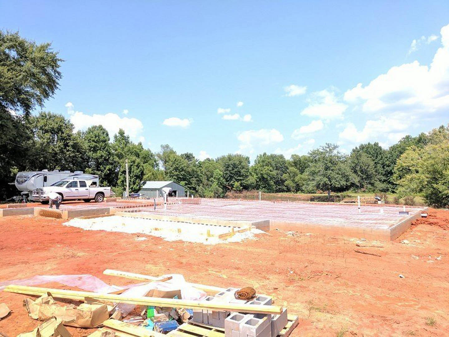 Framed custom home under construction with exposed wood and cardboard materials, white pickup truck parked on dirt lot, scattered bricks, trees and blue sky with clouds in
