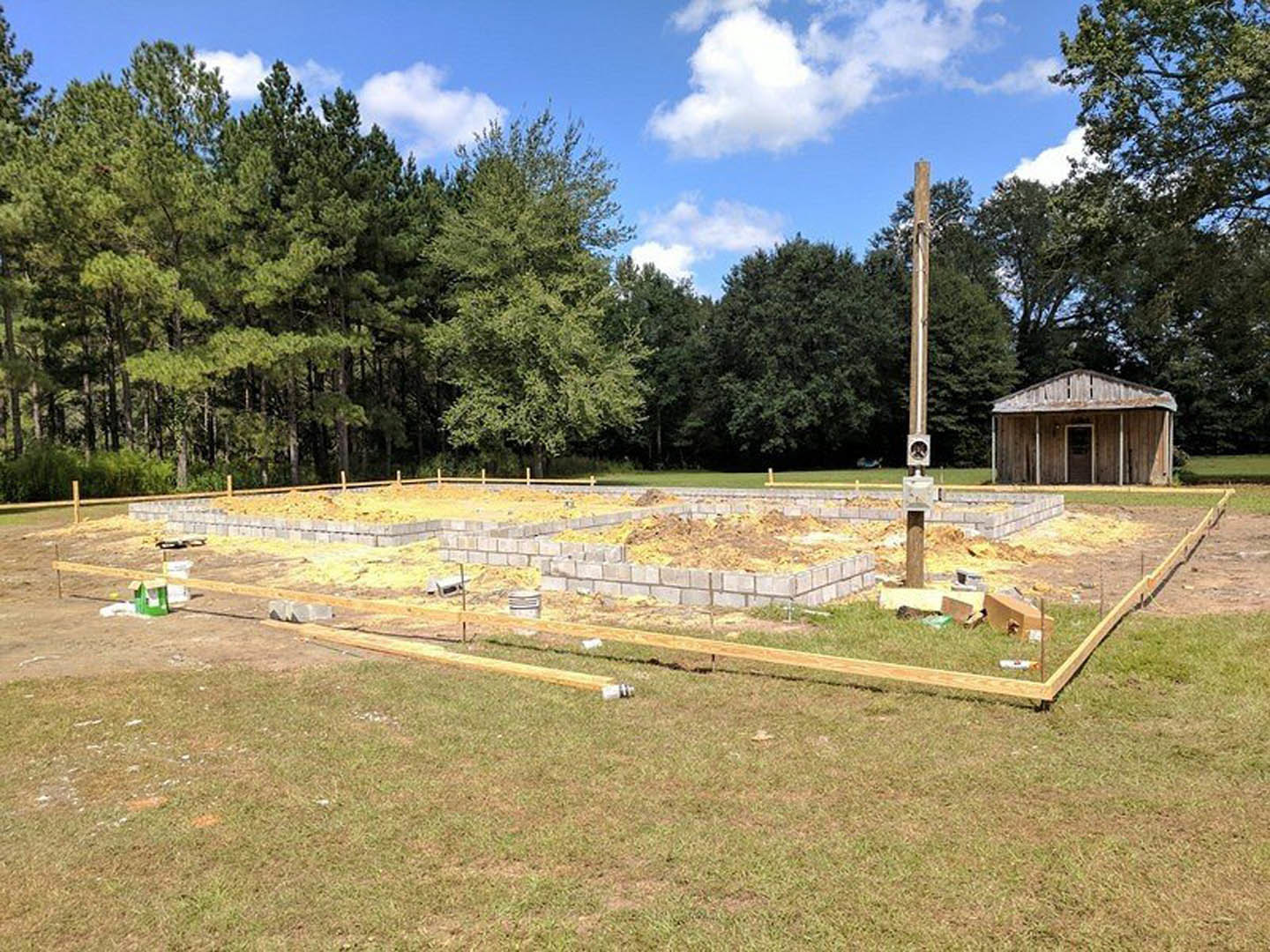 Wood-framed house under construction on grassy lot, surrounded by trees, with exposed foundation, piles of dirt and bricks, and a brown box in foreground beneath blue sky with