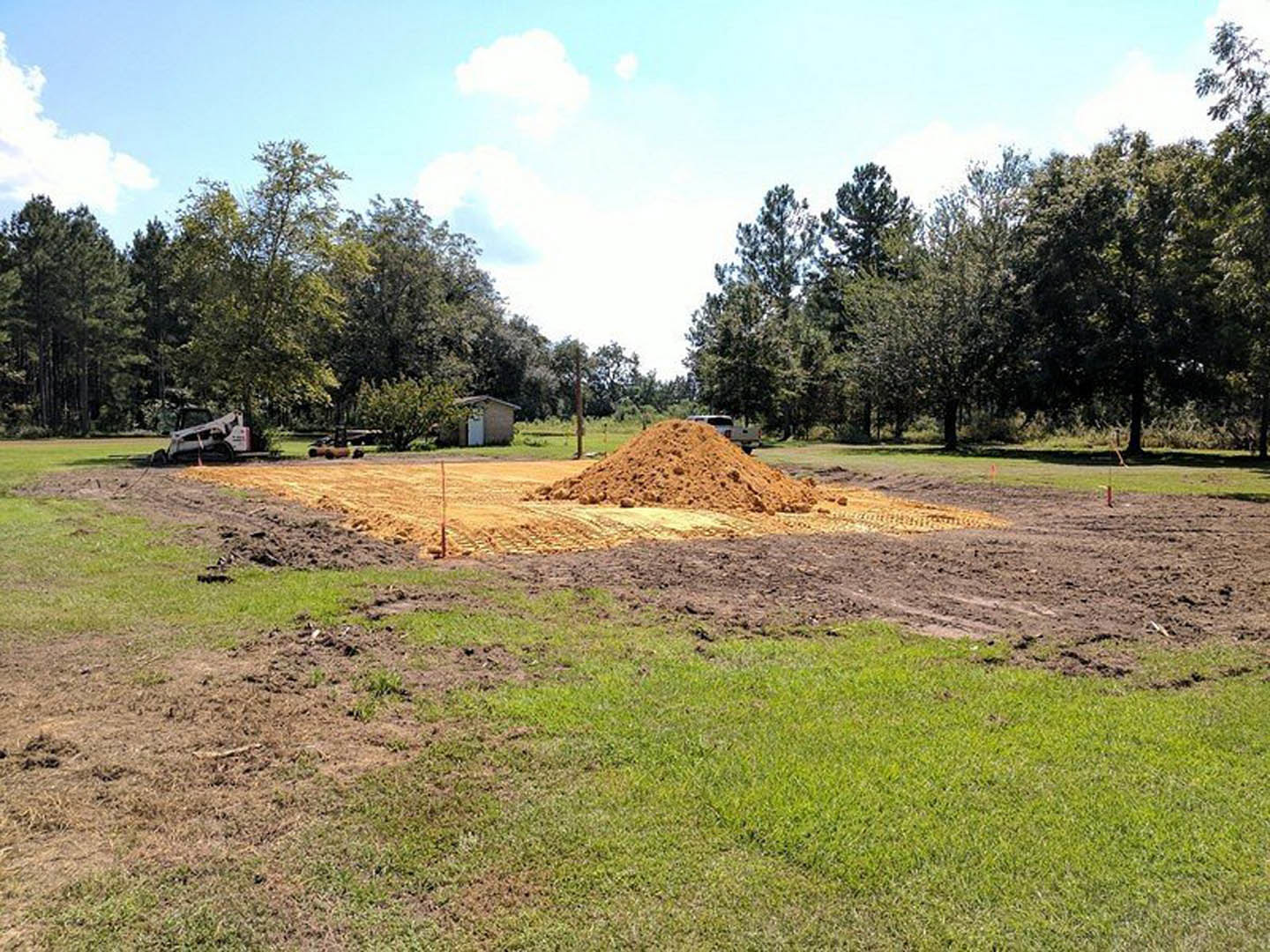 Mounded soil on grassy field with scattered trees, small structure with door, and blue sky with clouds