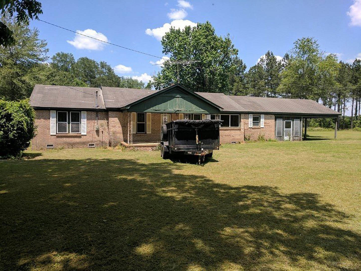 White two-story house with gray roof, black shutters, and large front windows; black trailer with cover parked on grassy lawn; mature trees and cloudy sky in background.