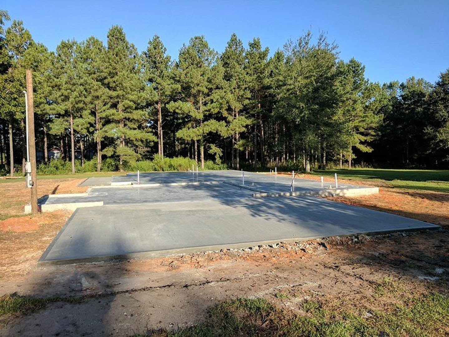 Smooth concrete slab foundation bordered by dirt and grass, tall trees and blue sky in background, partial shadow and metal pole visible in foreground