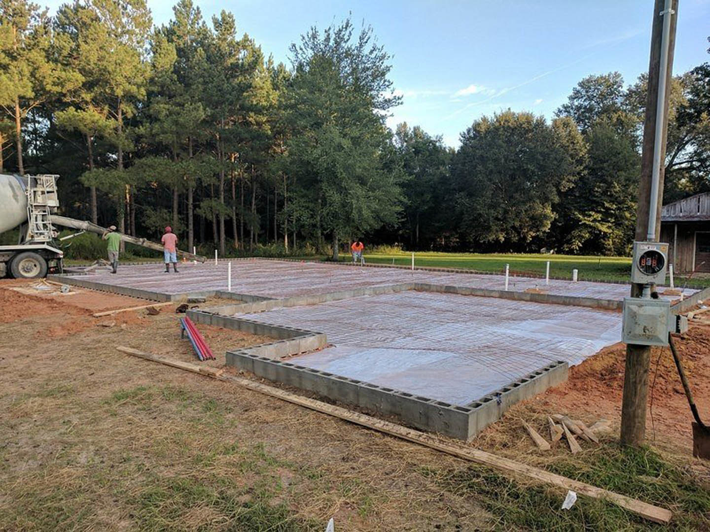 Concrete foundation slab surrounded by grass and trees, group of people standing on site, playground slide and ladder visible in background