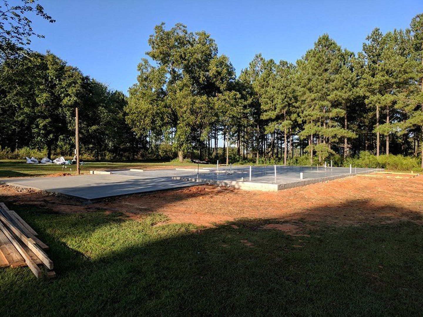 Concrete slab foundation surrounded by grass field, scattered wood planks, and mature trees in the background