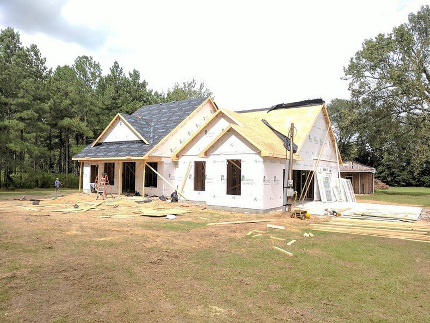 Partially built house framed with exposed wood beams, several workers on site, grassy lot with scattered construction materials, leafy tree in foreground, cloudy sky overhead